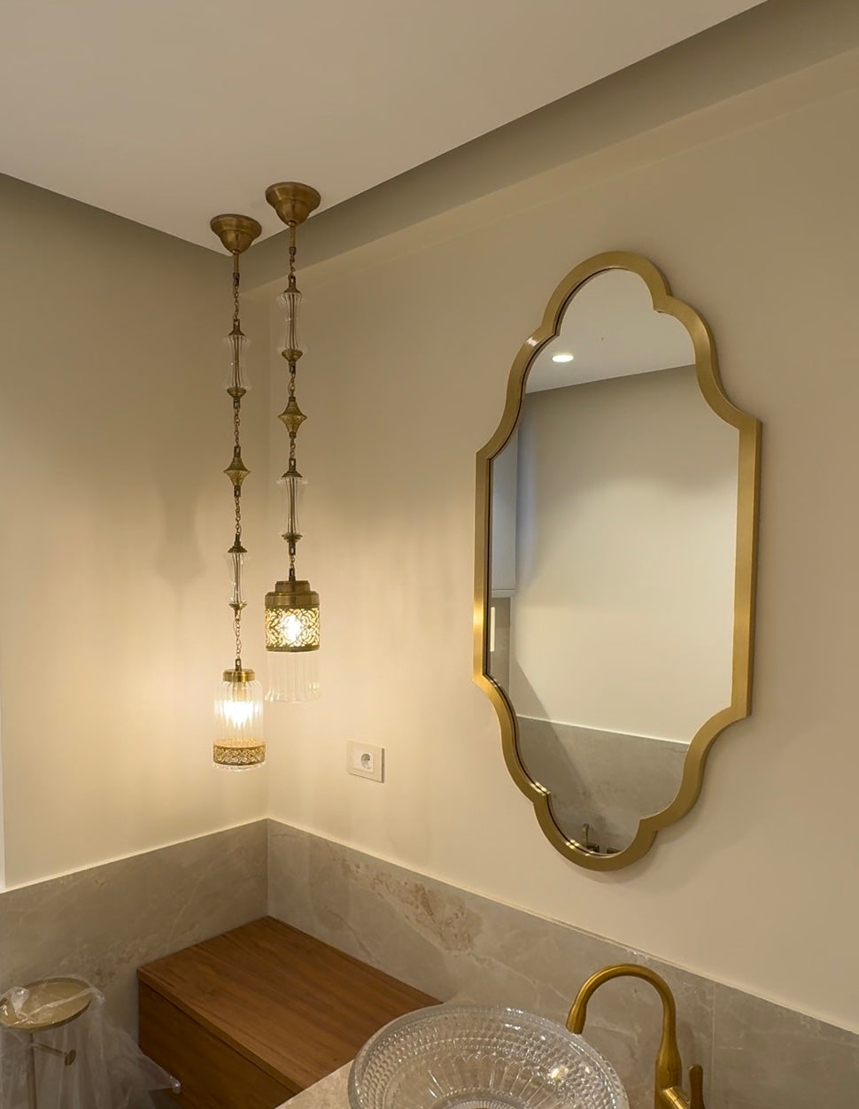 A bathroom interior featuring a large scalloped gold-framed wall mirror, two hanging brass pendant lights, a wood vanity bench, and a glass vessel sink with a gold faucet.